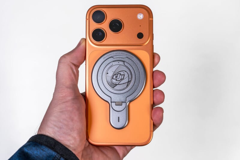 A hand holds an orange smartphone with three rear cameras, featuring a circular metallic DJI accessory attached to the back. The background is plain white.