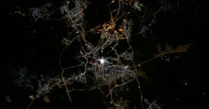 A city is illuminated at night, with bright lights outlining roads and neighborhoods, seen from above against a dark background. The city center appears brightest, with networks of lights branching outward.
