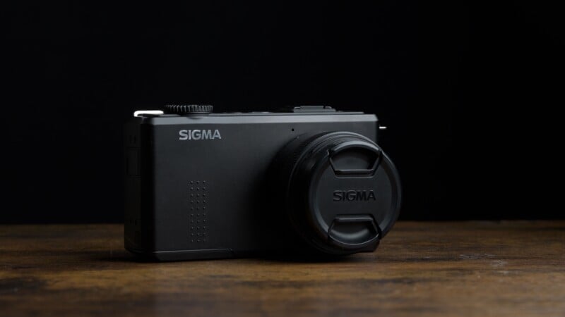 A black Sigma digital camera with a lens cap on, placed on a wooden surface against a dark background.