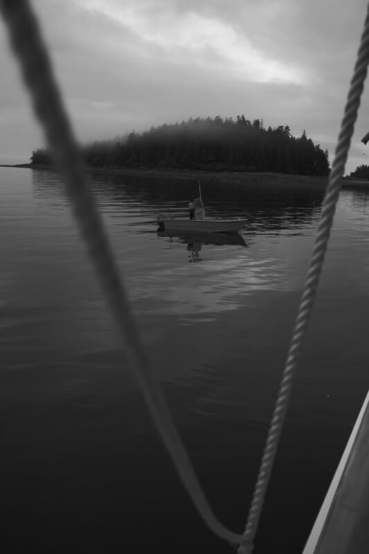 A small motorboat floats on calm water near a forested island, seen through two blurred rope lines in the foreground under a cloudy sky. The image is in black and white.