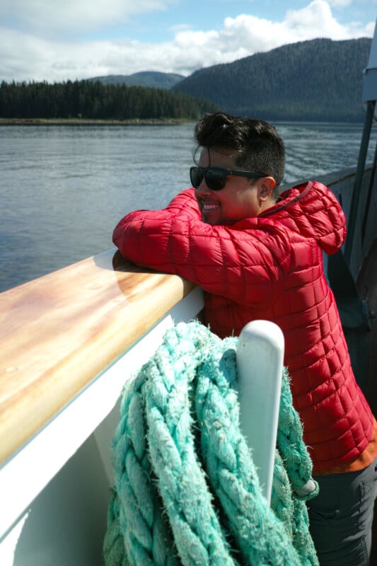 A person in a red jacket and sunglasses leans on the railing of a boat, smiling, with green rope nearby and a calm lake, forest, and mountains in the background under a partly cloudy sky.