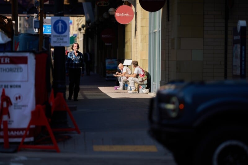 Two people sit on a sidewalk with belongings beside them, partially in sunlight. A person walks on the street toward the camera. A blue vehicle and red construction barriers are visible in the foreground.
