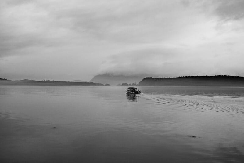 A small boat moves across a calm, foggy lake, leaving gentle ripples behind. Distant, tree-covered islands and hills are partially obscured by mist under a cloudy sky. The image is in black and white.