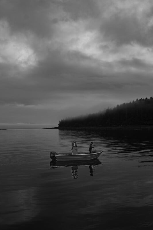 A lone person stands on a small motorboat floating on calm water under a cloudy, overcast sky, with a forested shoreline in the distance. The scene is in black and white, creating a moody and tranquil atmosphere.