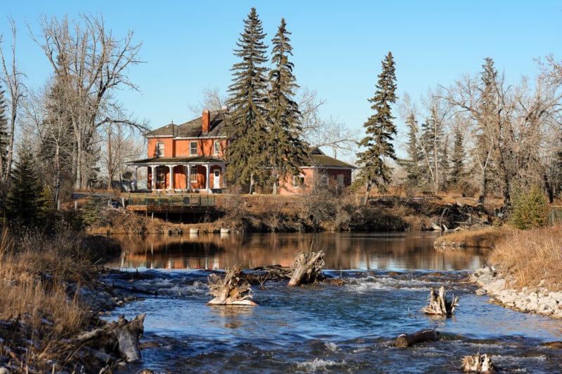 A large brick house with a wraparound porch sits near a calm river, surrounded by leafless trees and tall evergreens under a clear blue sky. Tree stumps and branches are visible in the shallow water in the foreground.