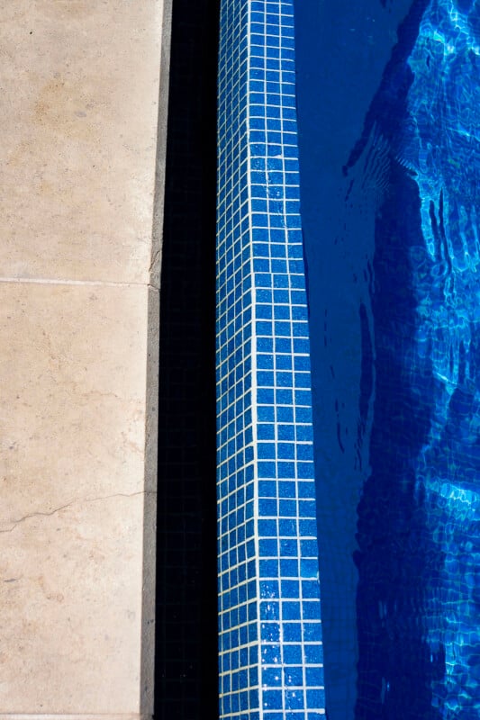 Close-up view of a swimming pool edge with blue mosaic tiles next to a beige stone pool deck. The water in the pool appears clear and vibrant blue, reflecting light.
