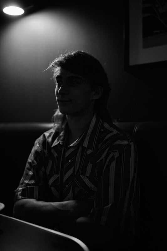 Black and white photo of a person with long hair sitting at a table in low light, arms crossed, wearing a striped shirt. A single overhead light casts shadows, leaving the person's face mostly in silhouette.