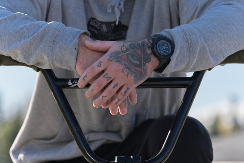 A person in a gray hoodie rests their tattooed hands, featuring an eye and symbols, on the handlebars of a BMX bike. They are wearing a black wristwatch. The background is blurred and outdoors.