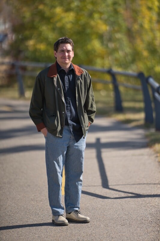 A man stands outdoors on a paved path with his hands in his pockets, wearing a green jacket, blue jeans, and light-colored shoes. Trees with green and yellow leaves and a metal railing line the background.