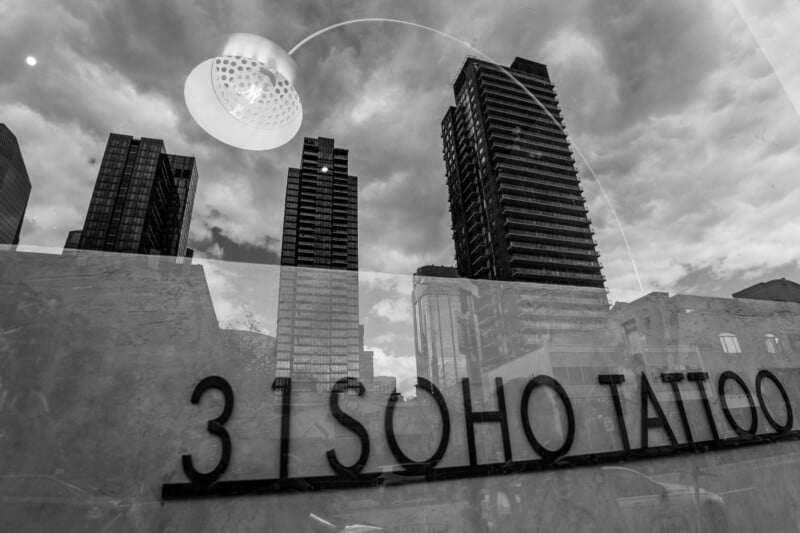 Black and white photo showing high-rise buildings reflected in glass. The words "31 SOHO TATTOO" appear on the glass, with a round ceiling light above and a cloudy sky in the background.