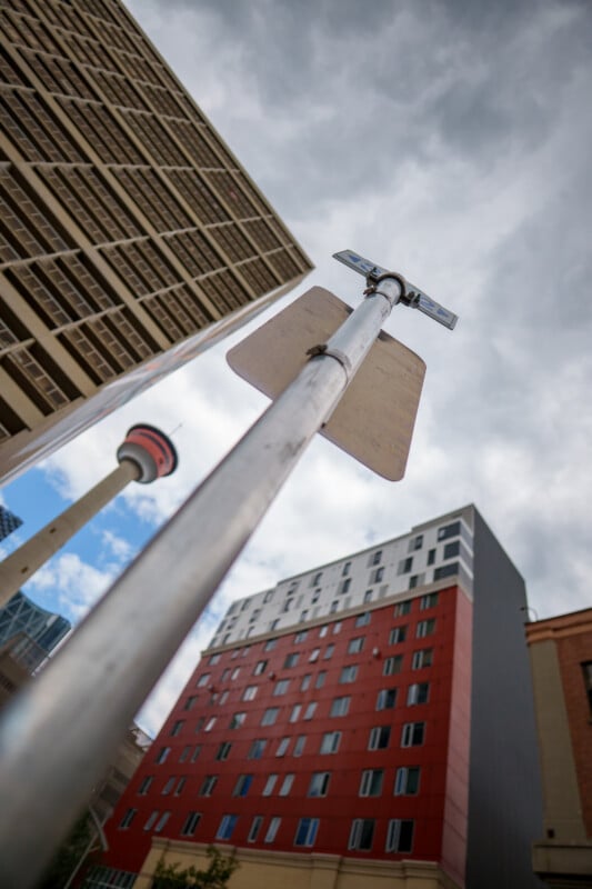 A low-angle view of a street signpost in front of tall buildings, including a red and white tower, against a cloudy sky. The perspective emphasizes the height of the signpost and surrounding architecture.