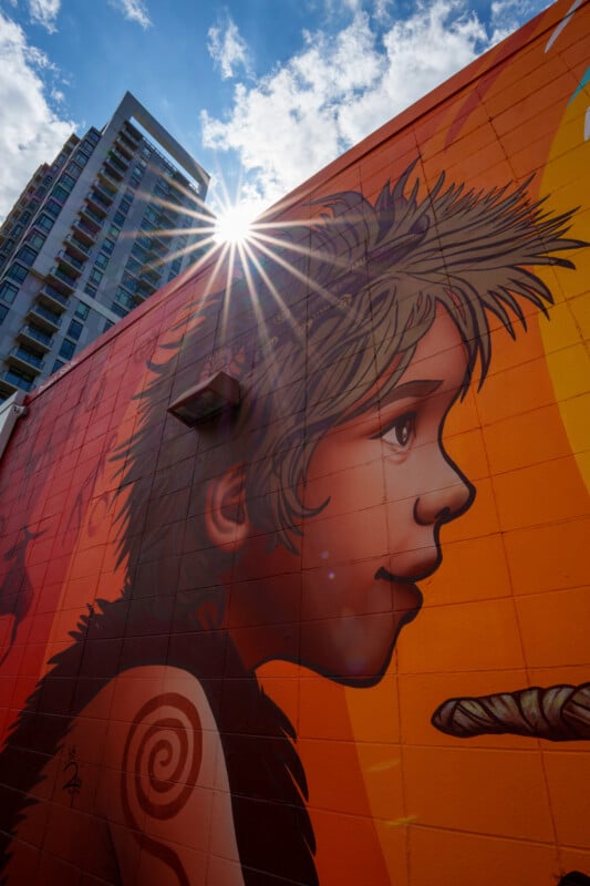 A vibrant mural of a child's face with spiky hair is painted on a building wall, with a bright sunburst overhead and a modern high-rise in the background under a partly cloudy sky.