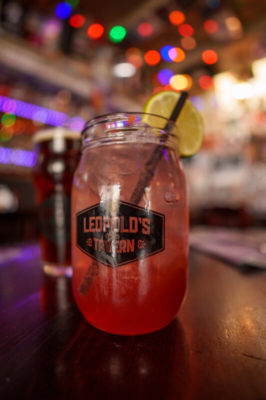 A pink cocktail with ice, a lime wedge, and a straw in a mason jar labeled "Leopold's Tavern," sits on a wooden table with colorful lights blurred in the background.