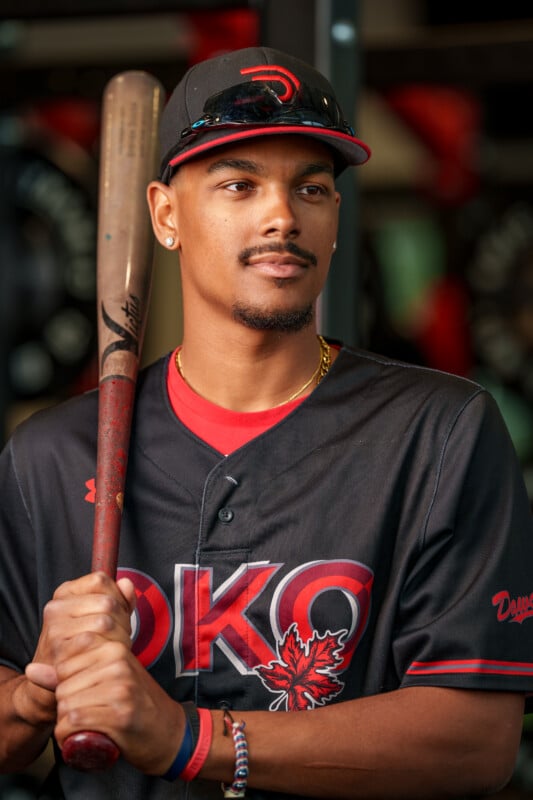 A baseball player wearing a black and red Okanagan jersey and cap holds a bat over his shoulder, looking confidently ahead. He wears a gold chain, bracelets, and stud earrings, with stadium seating blurred in the background.