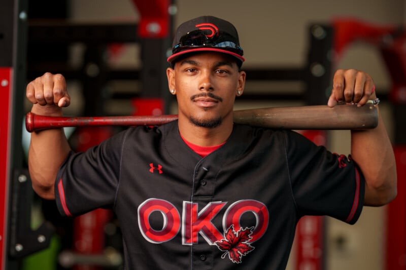 A baseball player in a black "OKO" jersey with a maple leaf design rests a bat on his shoulders, wearing a cap and sunglasses on his head, looking confidently at the camera.