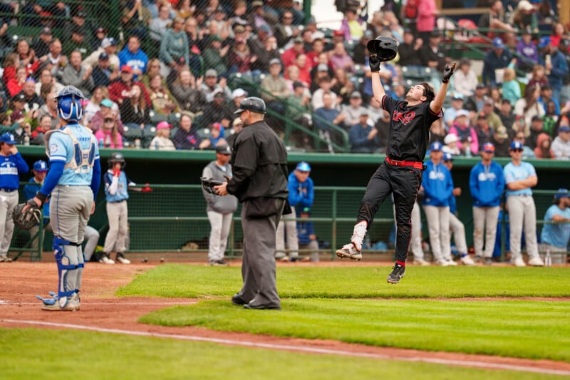 A baseball player in a black uniform leaps in celebration after scoring a run, holding his helmet up. An umpire, catcher, and crowd of spectators watch in the background.