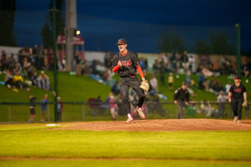 A baseball pitcher in a black and red uniform celebrates on the mound during a night game, with a blurred crowd and teammates in the background.