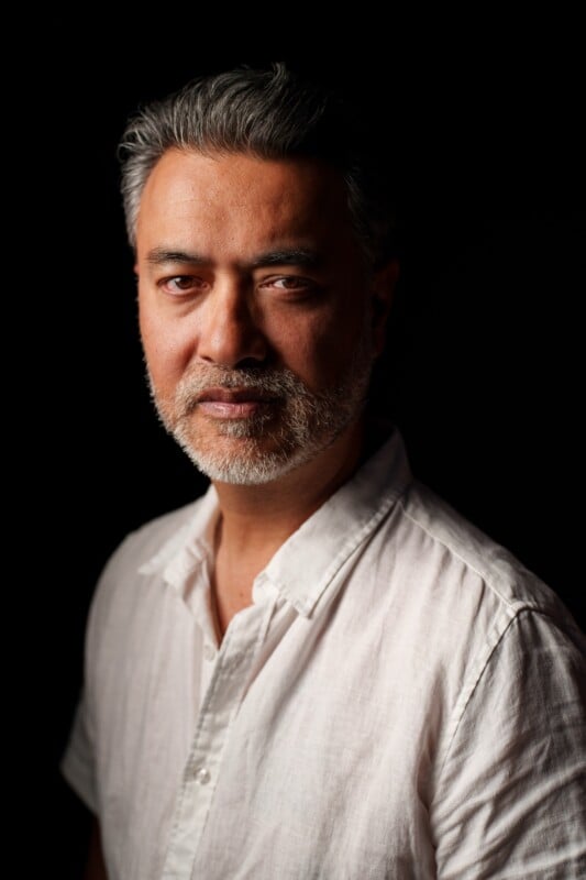A middle-aged man with gray hair and a beard, wearing a white button-up shirt, stands against a dark background, softly lit from the side.