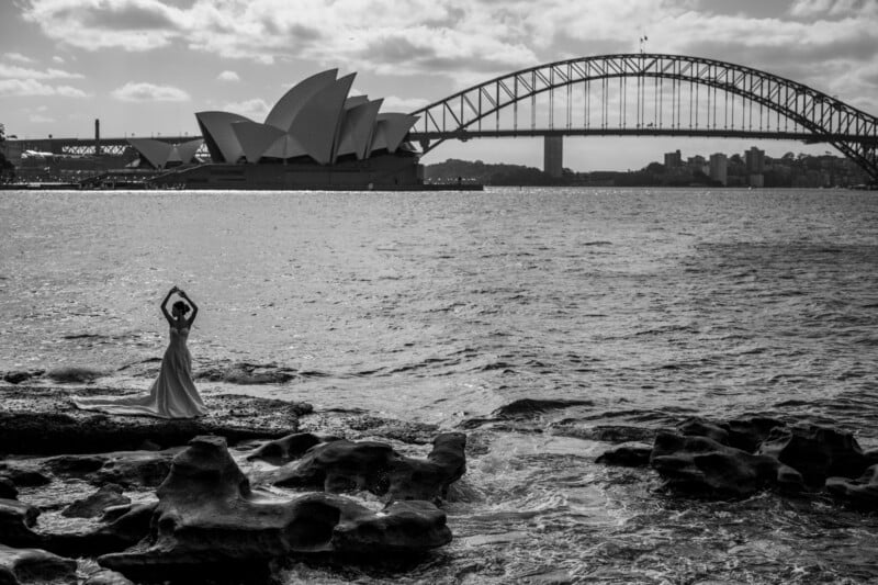 A person in a long dress stands on rocky shore with arms raised, facing the Sydney Opera House and Sydney Harbour Bridge across the water under a partly cloudy sky. Black and white photo.