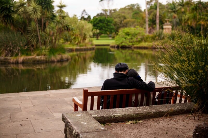 A couple sits closely together on a wooden bench, facing a tranquil pond surrounded by lush greenery in a park. The scene is peaceful and serene.