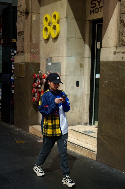 A person in a blue and yellow jacket, gray sweatpants, and a black cap walks on a city sidewalk while looking at their phone. Behind them is a building with the number 88 in yellow above the doorway.