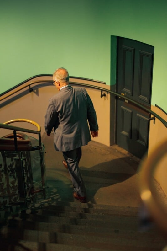 A man in a gray suit walks down a curved staircase with a green wall and a closed, dark-colored door in the background. The lighting is warm and soft, casting gentle shadows on the steps.
