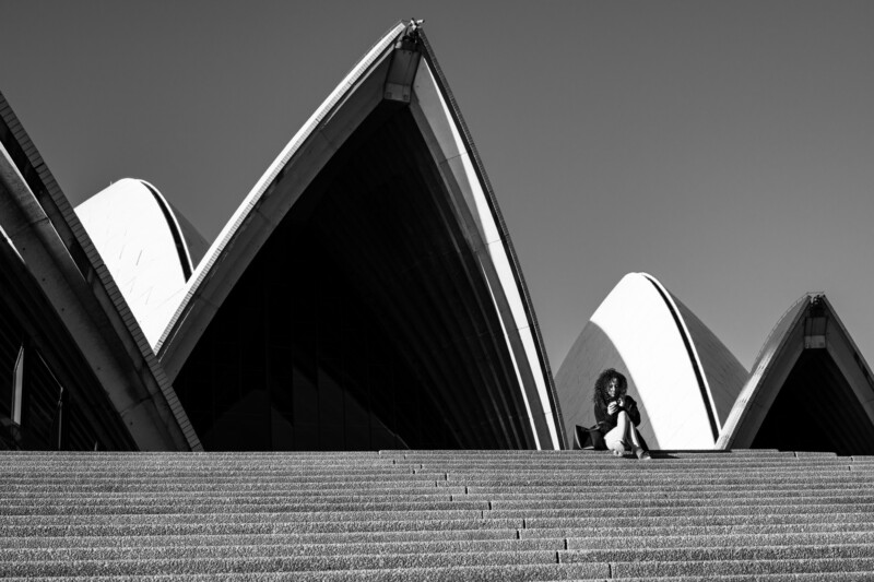 A person sits on the steps in front of the Sydney Opera House, with the building’s iconic sail-like roofs in the background under a clear sky. The photo is in black and white.
