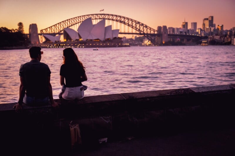 Two people sit on a stone ledge by the water at sunset, with the Sydney Opera House and Harbour Bridge visible in the background against a colorful sky.