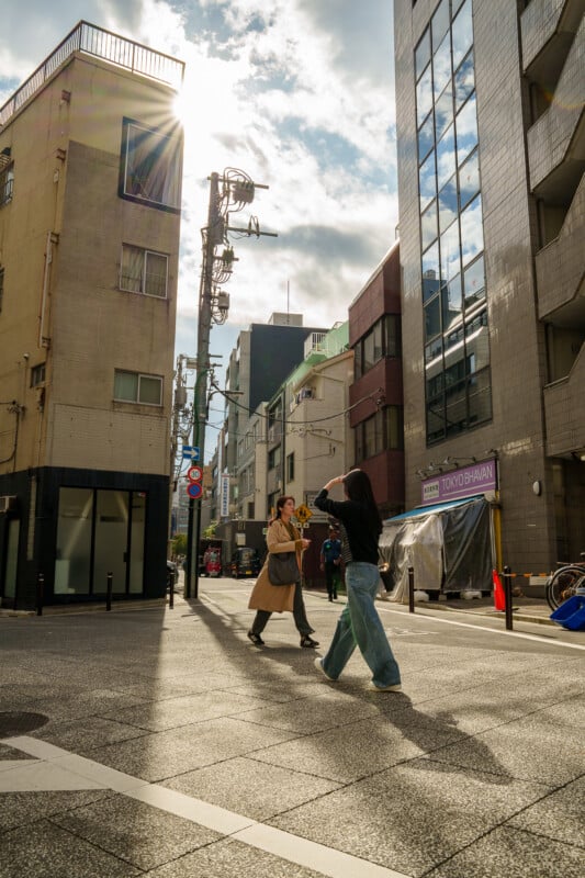 Two people walk and pose for photos on a sunlit city street, surrounded by tall modern and older buildings, with dramatic sunlight streaming through the clouds above.