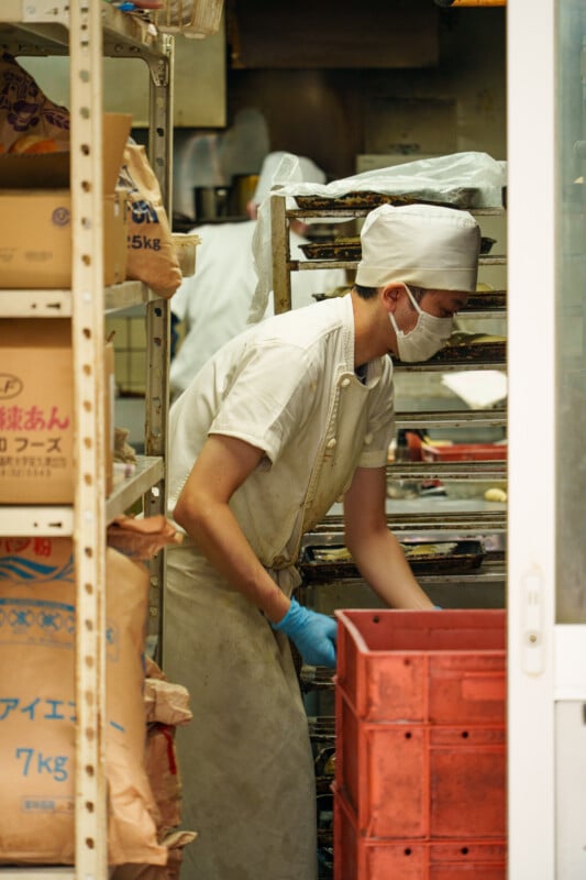 A baker wearing a white uniform, cap, and face mask arranges trays on a rack in a bakery kitchen, surrounded by shelves with bags of ingredients and red plastic crates.