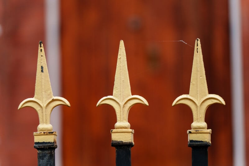 Close-up of three decorative yellow metal fence posts with fleur-de-lis tips, set against a blurred reddish-brown background. A faint spider web connects the posts.
