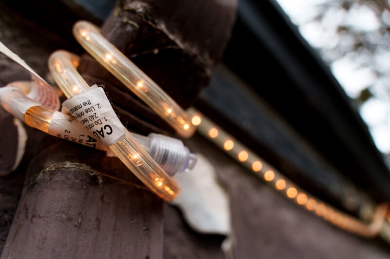 A close-up of an orange string light wrapped around a brown pipe on a wall, with a caution label visible and more lights blurred in the background.