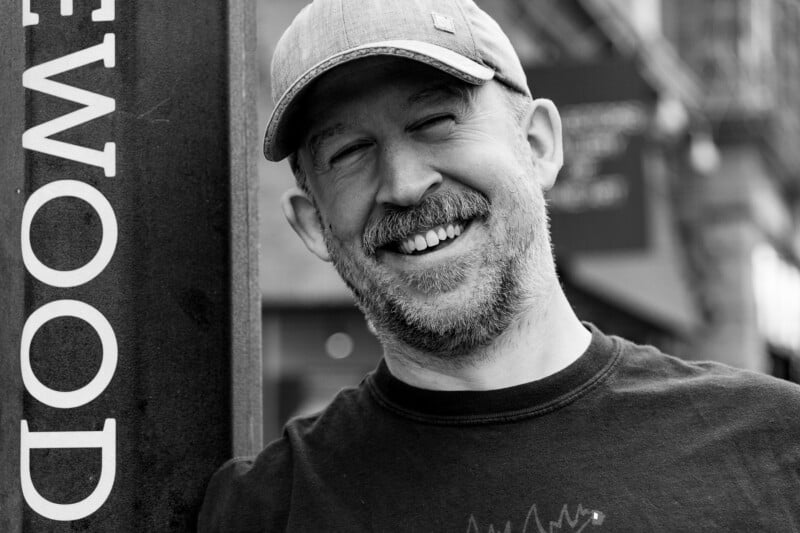 A bearded man in a cap smiles widely while standing outdoors next to a sign with partially visible text, in a black-and-white photo.