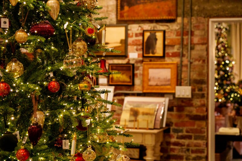 A decorated Christmas tree with gold and red ornaments and twinkling lights stands in a cozy room with brick walls, framed art, and a mirror reflecting the tree’s glow.