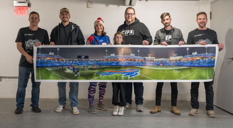 Six people stand indoors, smiling and holding a long panoramic photo of a football stadium. One person wears a festive hat and colorful leggings; others wear casual clothes. The scene is well-lit with a concrete floor and white walls.