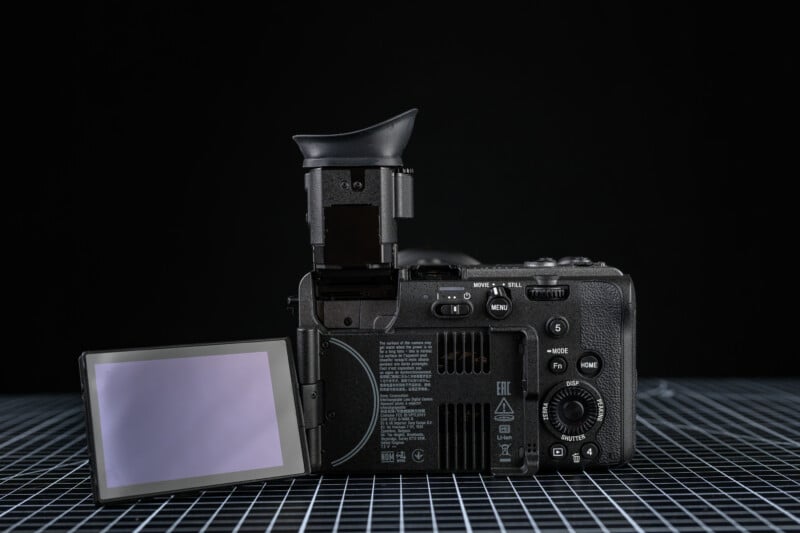 A digital camera with its LCD screen flipped out to the side and the electronic viewfinder raised, displayed on a grid-patterned surface against a black background.