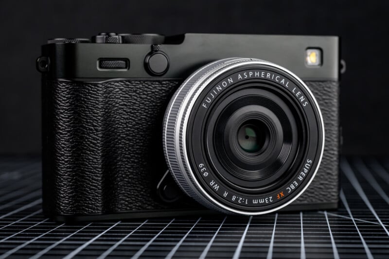 A close-up of a black Fujifilm digital camera with a Fujinon aspherical lens, placed on a black grid-patterned surface against a dark background.