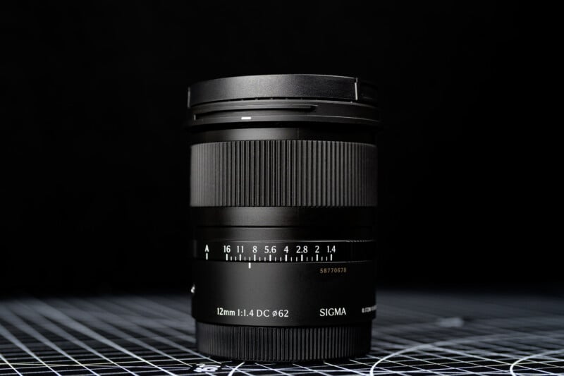 A Sigma 16mm f/1.4 DC DN camera lens is standing upright on a grid-patterned surface with a dark, blurred background. The lens markings and brand name are clearly visible.