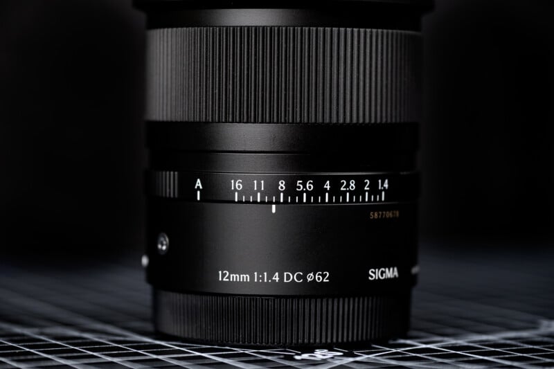 Close-up of a Sigma camera lens, showing the aperture ring with f-stop markings from 1.4 to 16, labeled "12mm 1:1.4 DC ø62." The background is blurred with a grid pattern beneath the lens.
