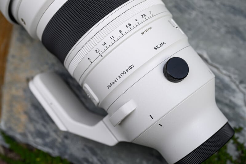 Close-up of a white Sigma 200mm F2 DG lens resting on a stone surface, showing the focus and aperture markings, Sigma branding, and part of the lens mount.