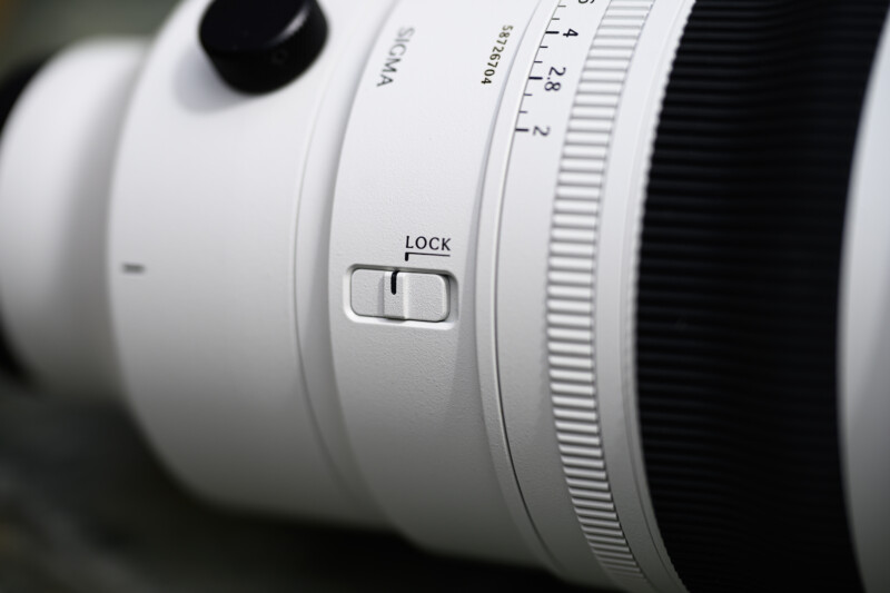 Close-up of a white Sigma camera lens showing a lock switch, focus and zoom rings, and engraved markings for aperture and model information. The texture and details of the lens are clearly visible.