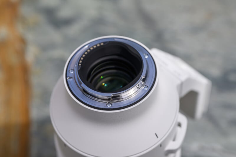 Close-up of the metal lens mount and rear glass element of a white camera lens, showing electronic contacts, with a blurred background.