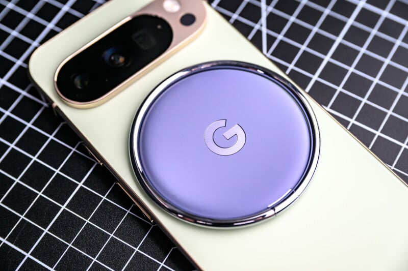 A close-up of a smartphone with a pale yellow back and a large black camera module, featuring a circular, purple Google-branded accessory attached to the back, resting on a black and white grid-patterned surface.