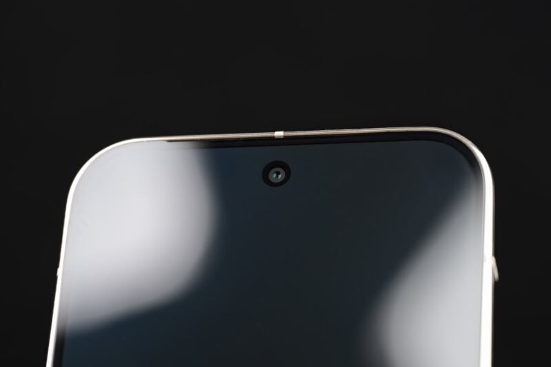 Close-up of the top portion of a smartphone against a black background, showing the front-facing camera centered above the screen. The screen reflects some light, creating white blurred shapes.