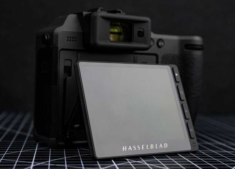 A close-up of a Hasselblad camera with its rear LCD screen flipped out, displaying the brand name. The camera is placed on a grid-patterned surface with a dark background.