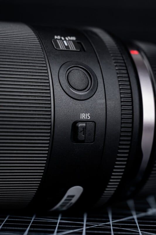 Close-up of a camera lens showing a ridged focus ring, a circular button, an AF/MF switch, and an IRIS switch on the lens barrel. The background is out of focus with a grid pattern beneath the lens.