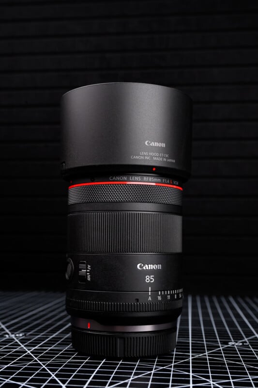 A Canon RF 85mm F1.2L camera lens with a lens hood is standing upright on a black-and-white grid surface, against a dark background with horizontal lines.