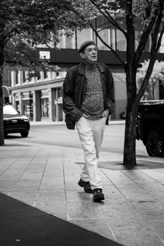 Black and white photo of an older man walking on a city sidewalk, wearing a cap, jacket, turtleneck sweater, and light pants. Trees and parked cars line the street in the background.