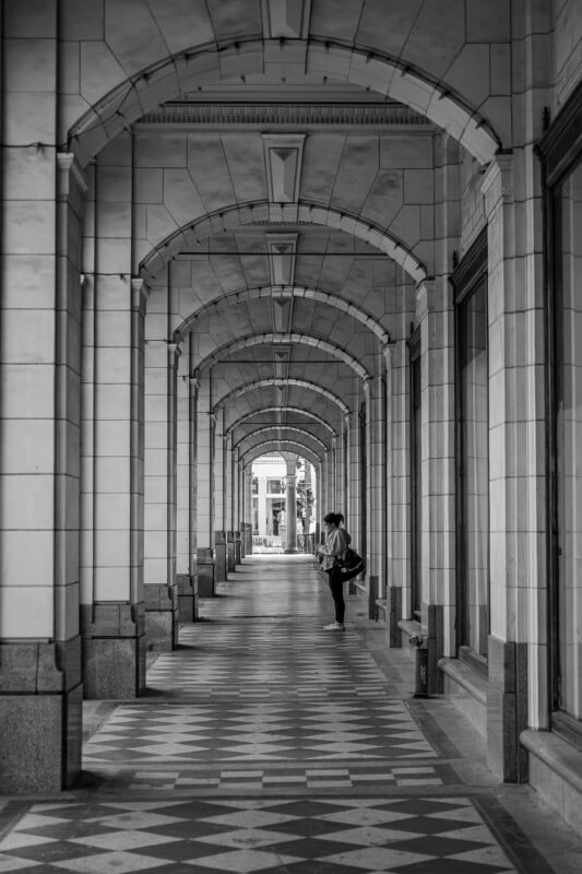 Black and white photo of a person standing in a covered walkway with arched ceilings and checkered floor tiles, surrounded by tall columns that create a sense of depth and symmetry.