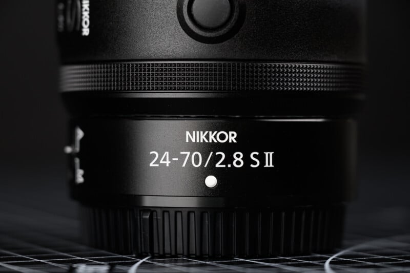 Close-up of a black NIKKOR camera lens, showing “24-70/2.8 S II” text prominently on the lens barrel, with focus on the branding and model details. The background is dark and out of focus.